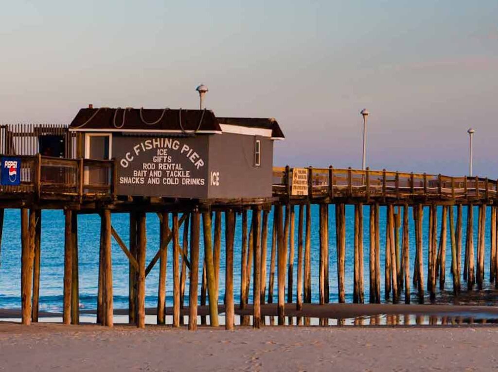 A wooden fishing pier with a small shack above the water, advertising Fishing supplies and snacks, extends over the ocean near a sandy beach.