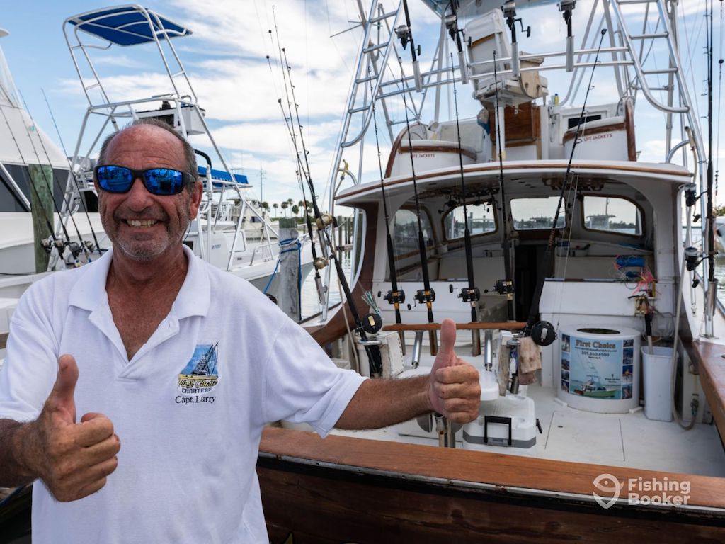 A man in a white polo shirt smiling and giving two thumbs up in front of a Blue Star fishing boat that's equipped with rods and gear.