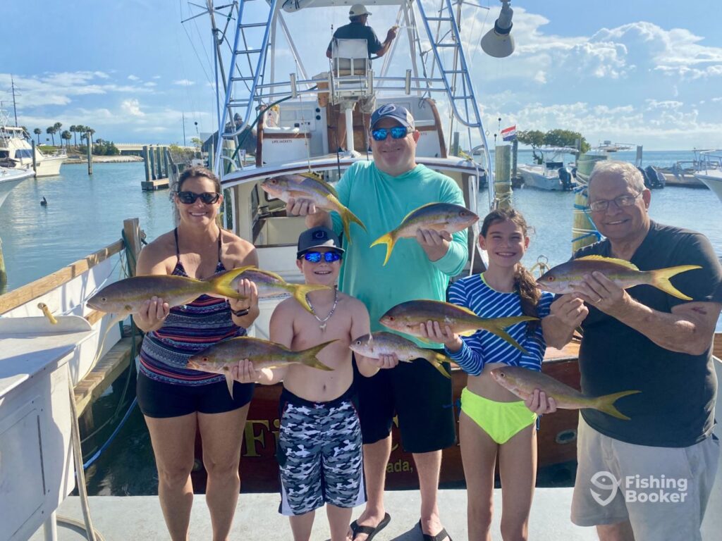 Three adults and two children stand on a dock, each holding Yellowtail Snapper, in front of a Blue Star fishing boat at a marina on a sunny day.