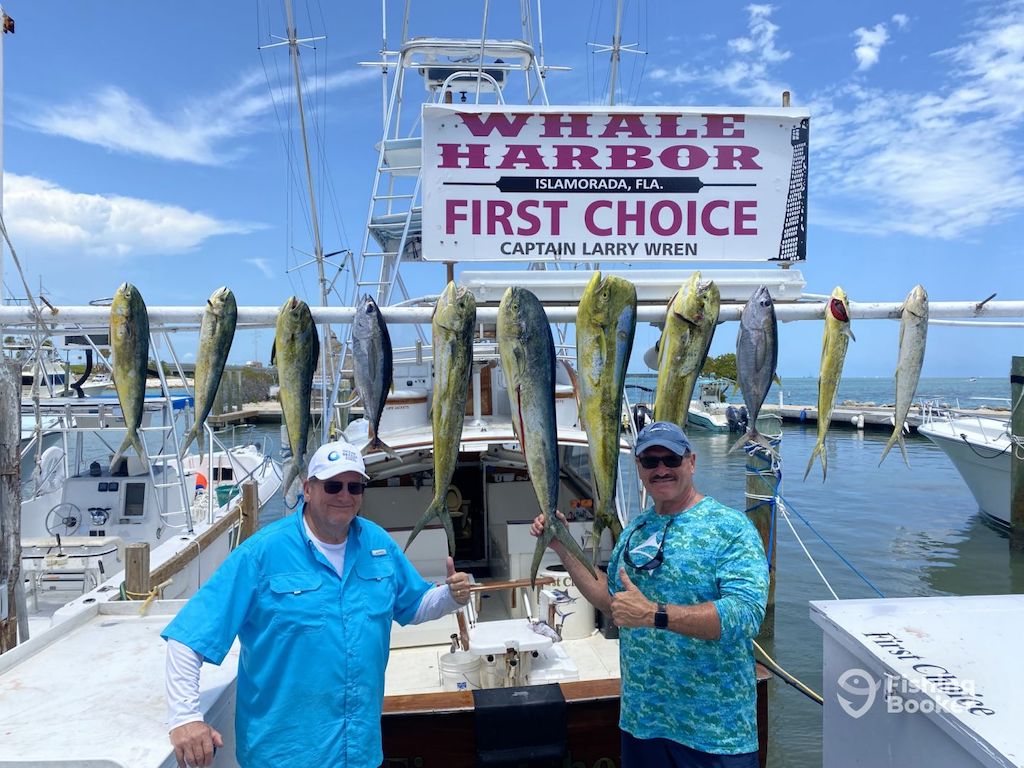 Two people are standing on a dock in front of a boat and a sign in Whale Harbor, Islamorada. Behind them, several fish, including numerous Mahi Mahi are hanging on display.