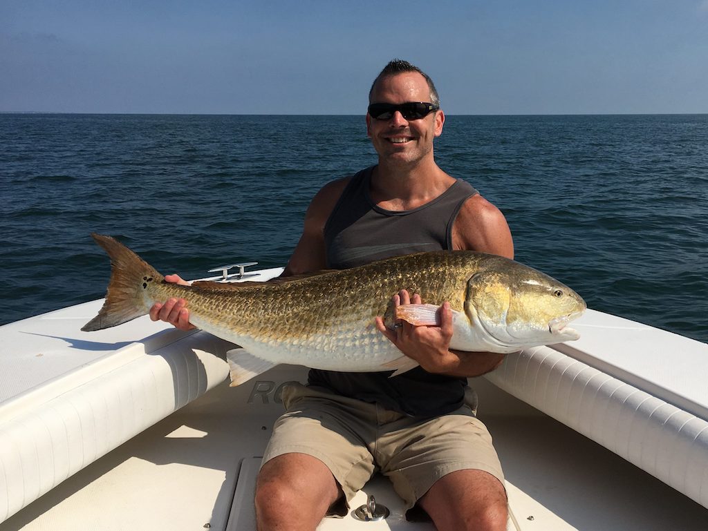 A man in a tank top and shorts's sitting on a boat in North Carolina, holding a large Redfish with the ocean in the background.