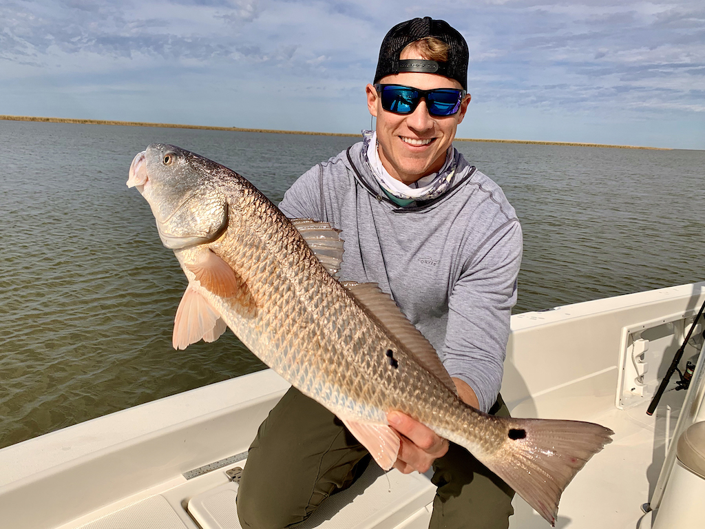 A man wearing sunglasses and a cap, holding a large Redfish on a boat with water and sky behind them.