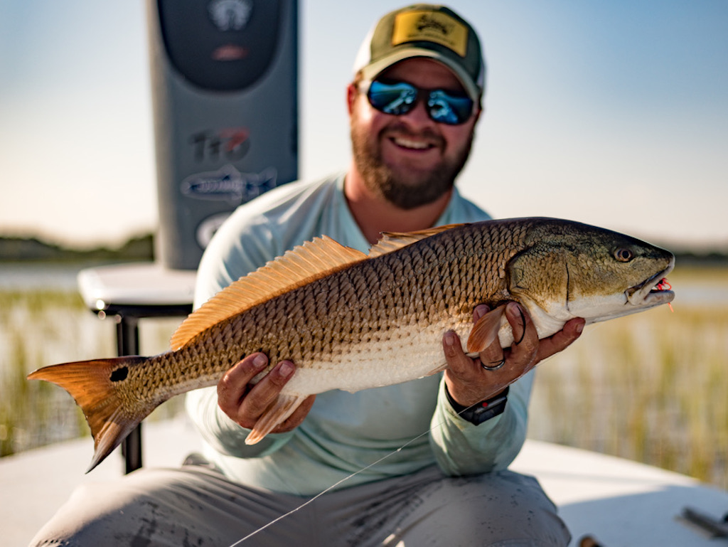 A man wearing sunglasses and a cap as he holds a large Redfish on a boat, with marshland in the background.
