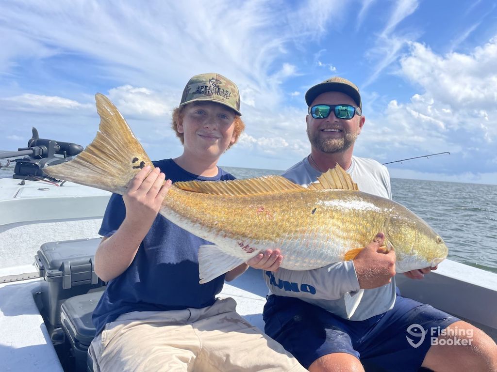 Two people are sitting on a boat, smiling and holding a large Redfish; open water and a partly cloudy sky are in the background.