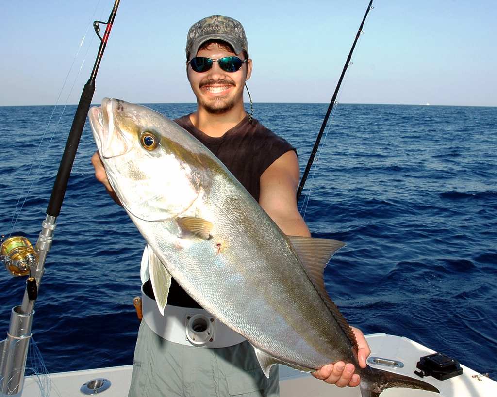 A man in a cap and sunglasses smiles while holding a large Reef Donkey on a boat, with fishing rods and the ocean in the background.