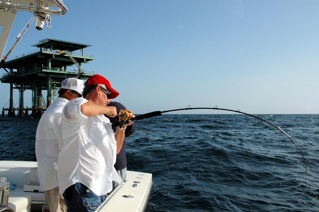 Three people on a boat are fishing in open water near an offshore oil platform; one person's battling a Reef Donkey with a bent fishing rod.