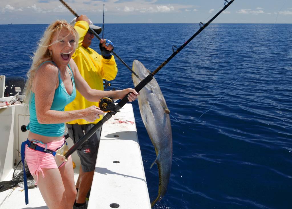 Two people on a boat are fishing; one’s reeling in a large Reef Donkey while the other helps, with the ocean and blue sky in the background.