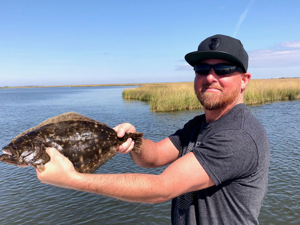 A man wearing sunglasses and a cap, holding a Flounder fish in both hands by the water. There are reeds behind him and a blue sky above.