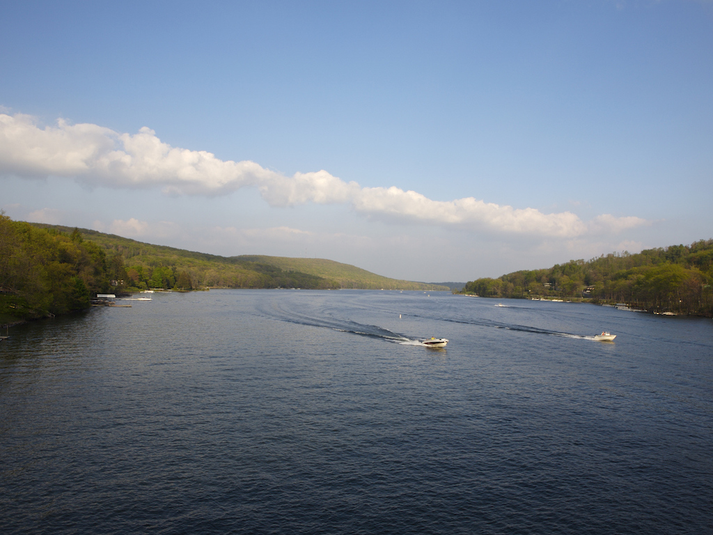 A wide lake with two motorboats making wakes, surrounded by green hills and under a blue sky.