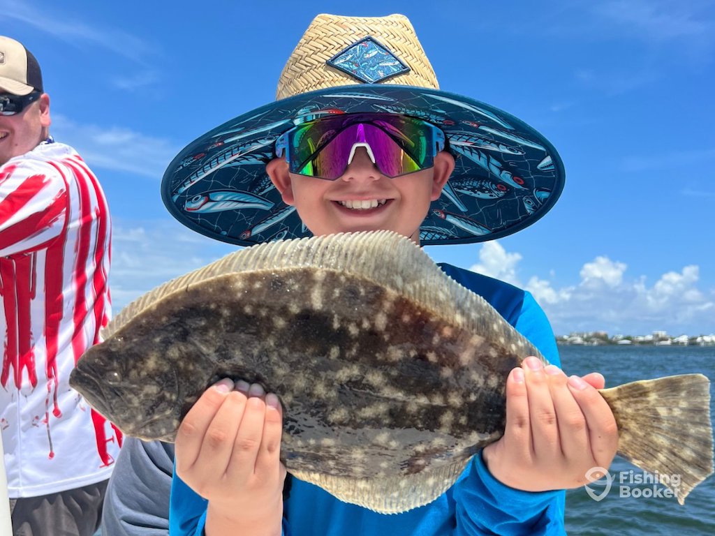 A smiling child in sunglasses and a hat holding a large Flounder with both hands on a boat, beneath a sunny blue sky.