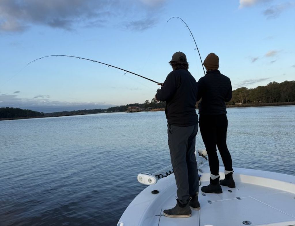 Two people in jackets and hats stand on a boat, fishing with rods over calm water under a mostly clear sky.