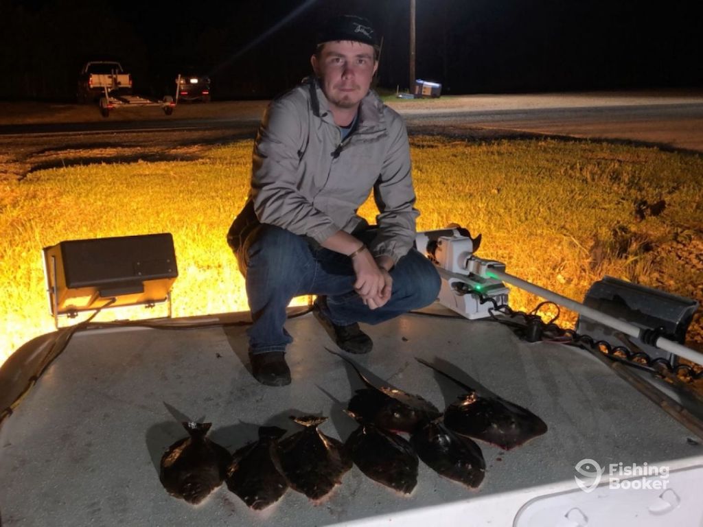 A man kneeling on a boat beside six Flatfish arranged in a row at night. There's a streetlight and a trailer in the background.
