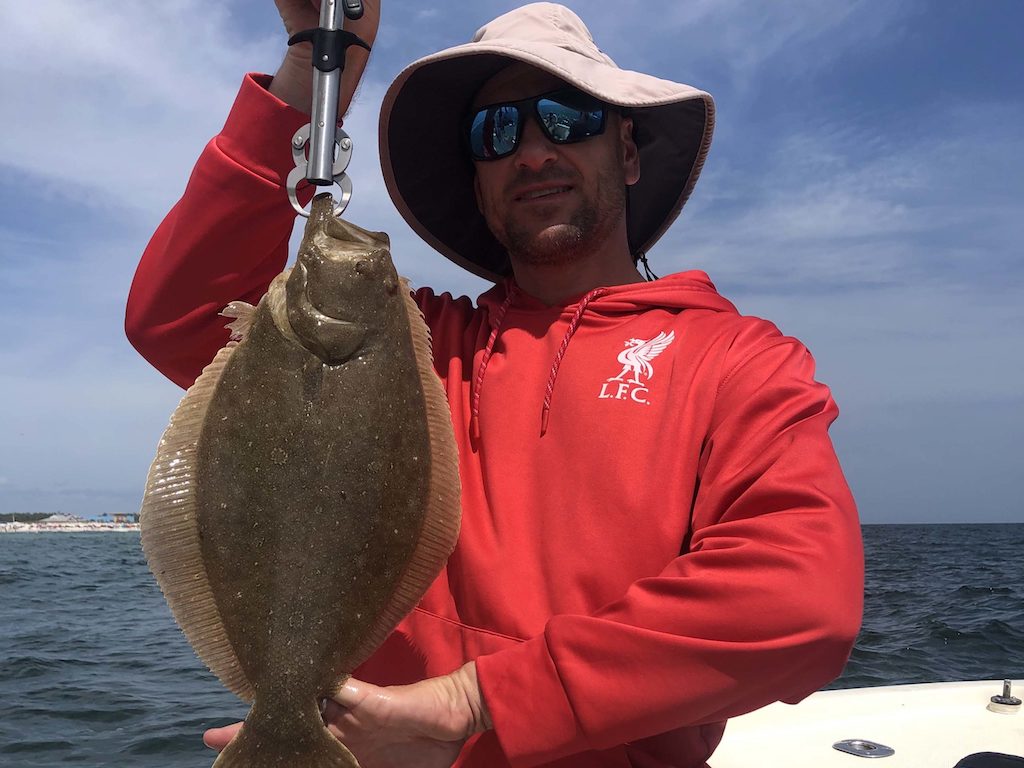A man in a red Liverpool FC hoodie holds up a Flounder with a clamp on a boat, with the ocean and sky behind him.