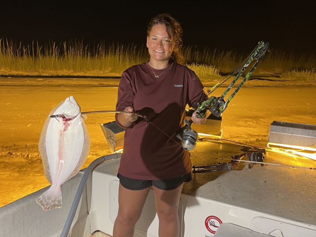 A woman standing on a boat at night, holding a fishing rod in one hand and a flat, white Flounder with a hook in the other.