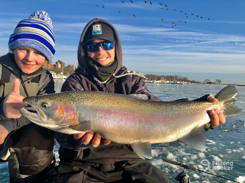 Two people in winter clothing hold a large fish, possibly a Steelhead, outside on ice during Wisconsin Trout seasons; there's a cityscape and flock of birds behind them.
