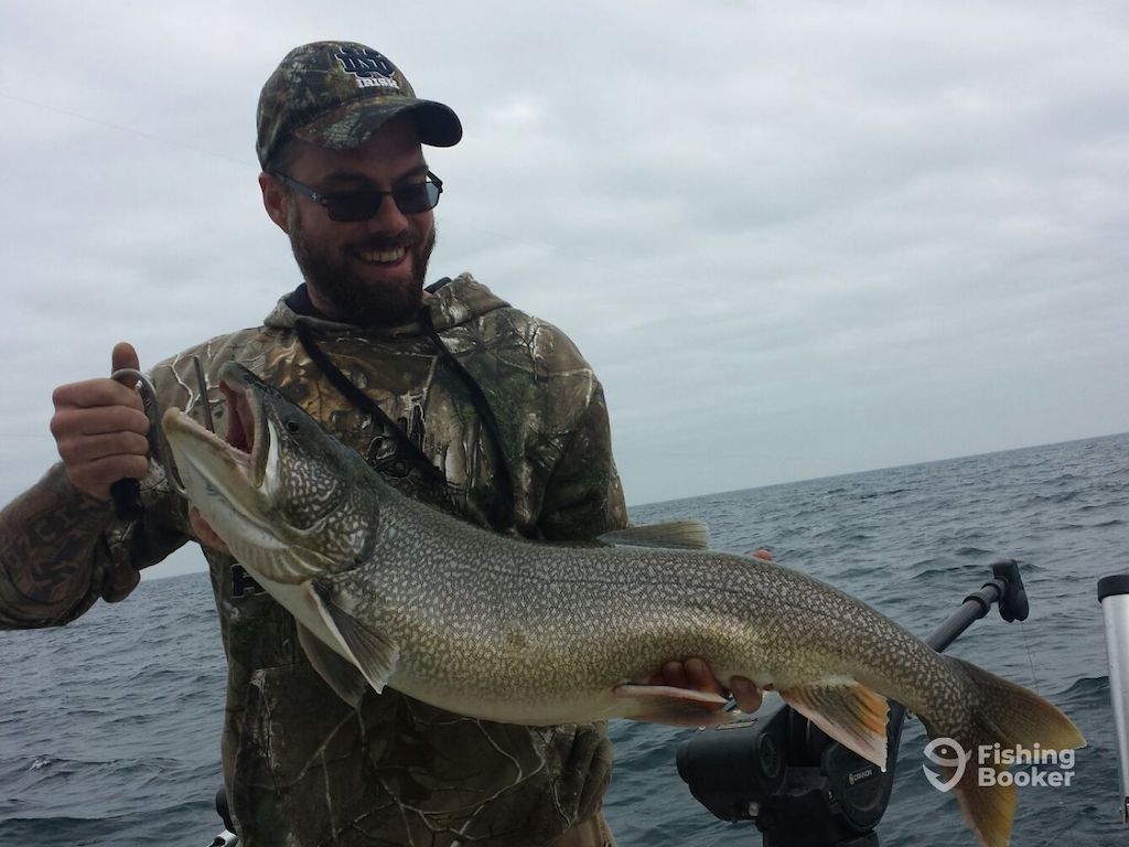 A man in camouflage clothing's holding a large Trout on a boat, celebrating Wisconsin Trout seasons with the ocean in the background.