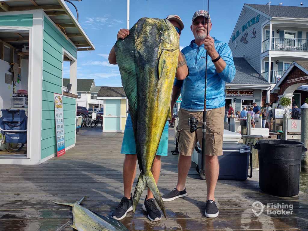 Two anglers pose on a dock holding a large Mahi-Mahi caught on their offshore trip in Ocean City in Maryland, with marina buildings and fishing gear in the background.