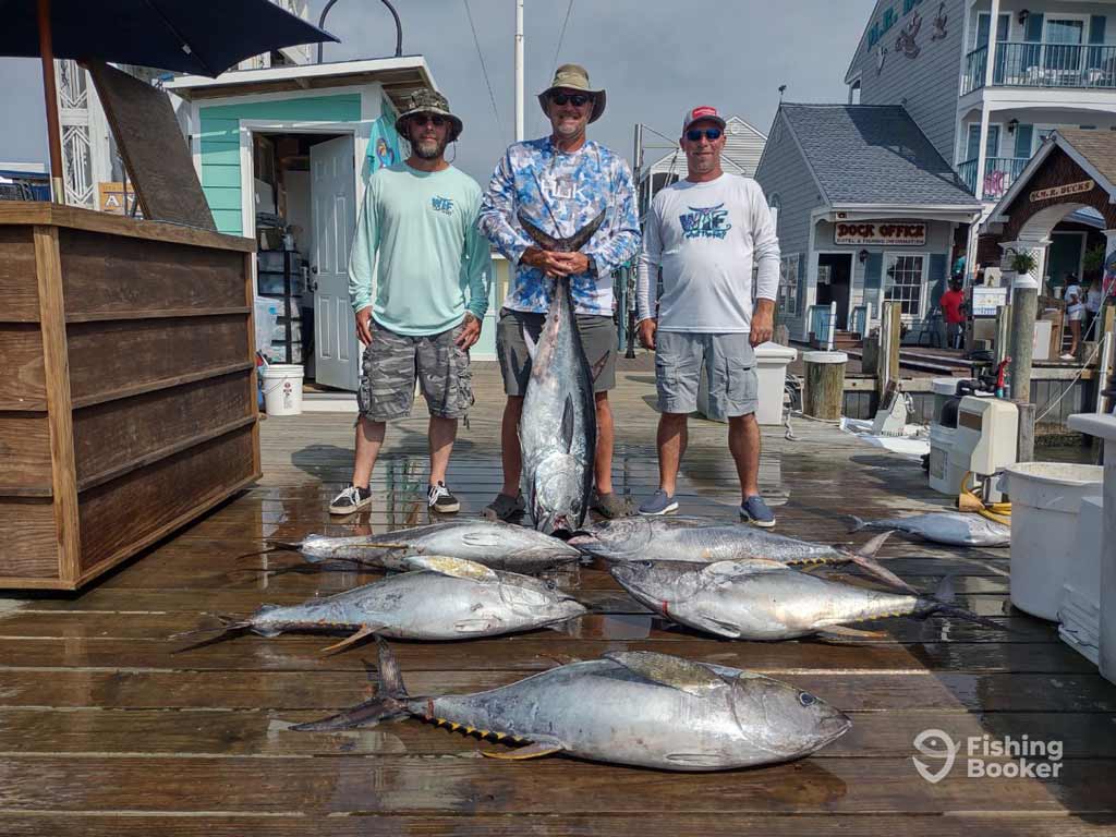 Three anglers stand on a dock behind their Tuna catches and one fish being held upright. There’re buildings and a dock office in the background.