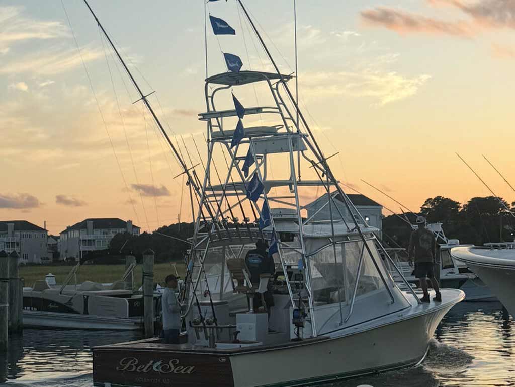 A white charter fishing boat called “BetSea” with blue Marlin flags is docked at sunset during the White Marlin Open. There are people onboard and houses in the background.
