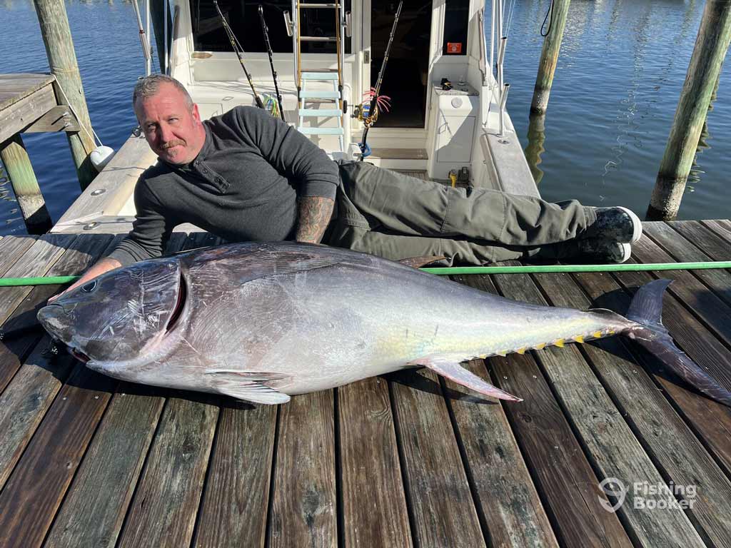 An angler’s lying on a wooden Manteo dock in North Carolina next to a huge Tuna, with a boat and water behind him.