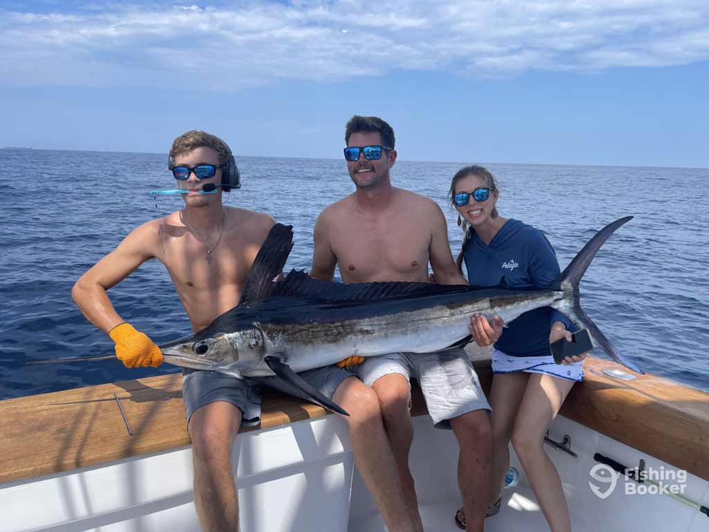 Three Billfish anglers are sitting on a boat and holding a large Marlin, with the ocean in the background. One person’s wearing gloves and sunglasses.
