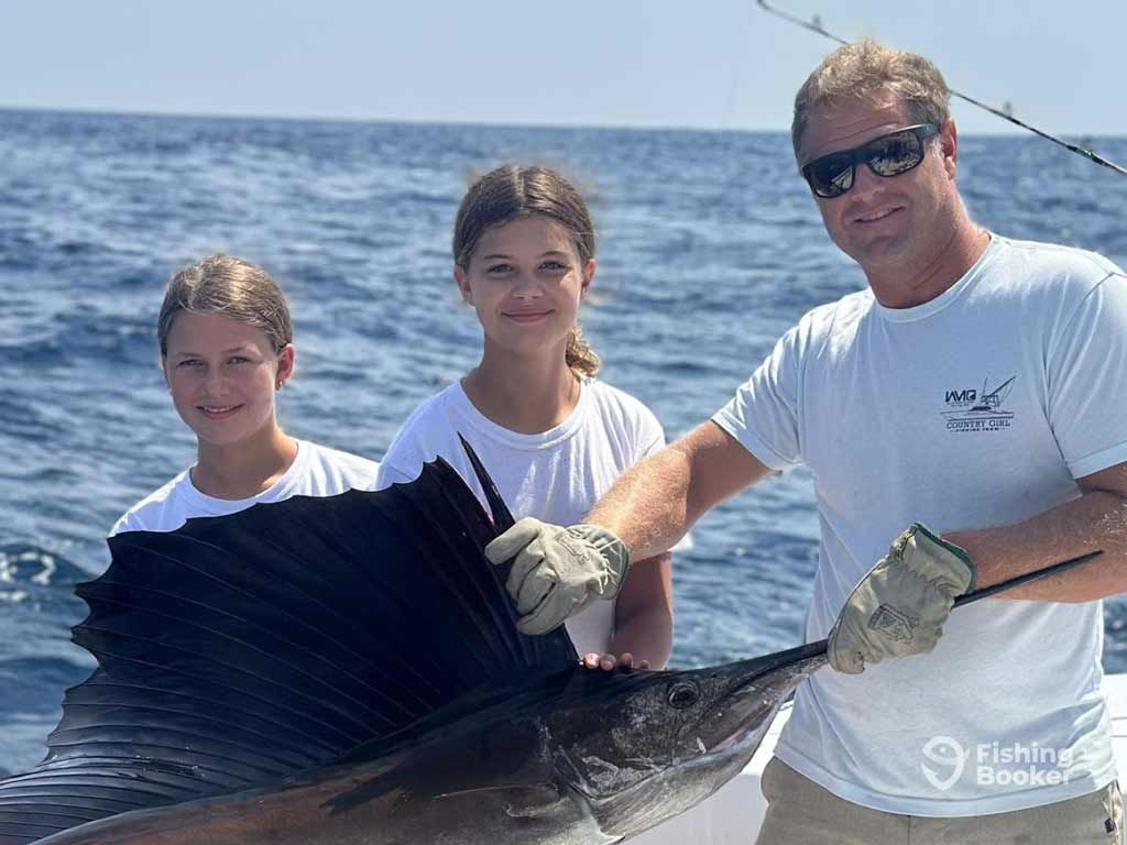 Three people, one adult and experienced angler with two kids, are on a boat and they are holding a large Sailfish; the ocean and a fishing rod are visible in the background.
