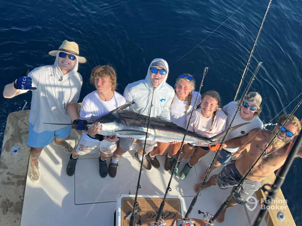 Seven anglers are posing on a deep sea fishing boat while holding a large Billfish behind the fishing rods, surrounded by bluewater.