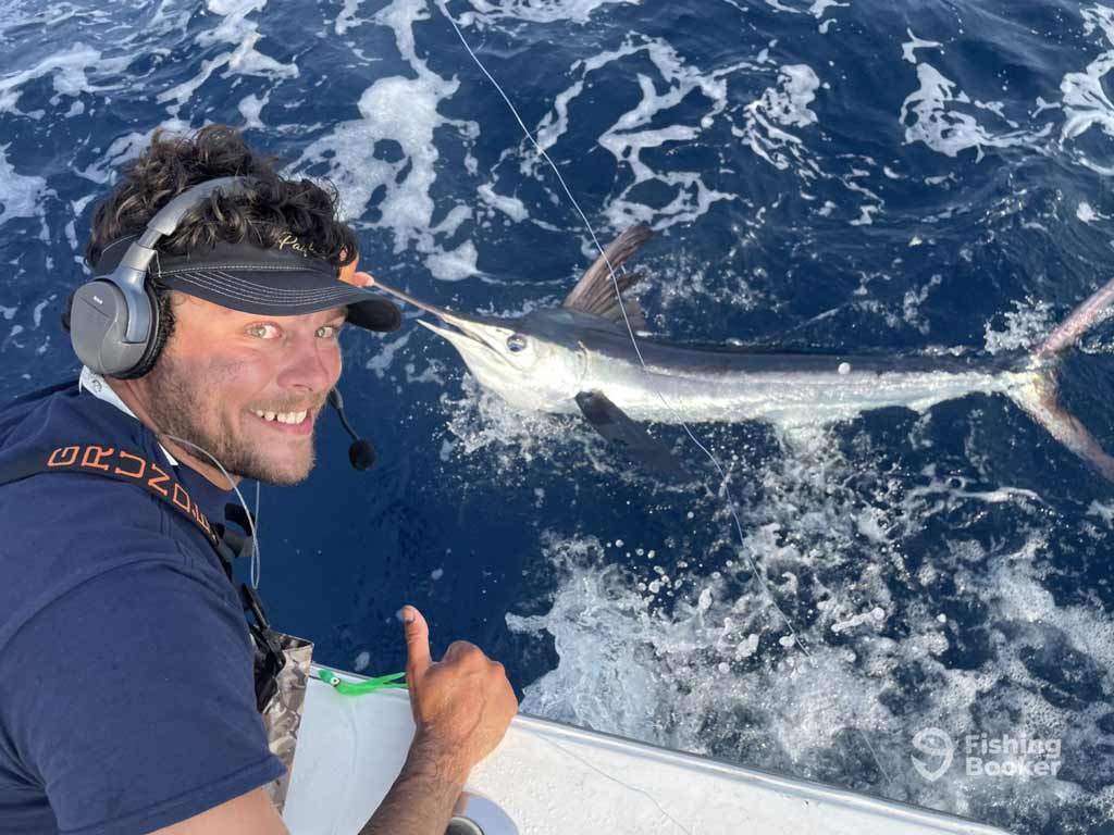 A charter captain in a headset’s giving a thumbs up while fishing on a boat at the Virginia Beach Billfish Tournament, holding a line attached to a large Marlin in the ocean.