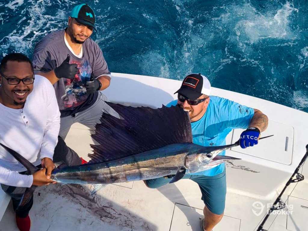 Three proud anglers on a boat hold a large Sailfish above the deck, a catch worthy of the Virginia Beach Billfish Tournament, while ocean water shimmers behind them.