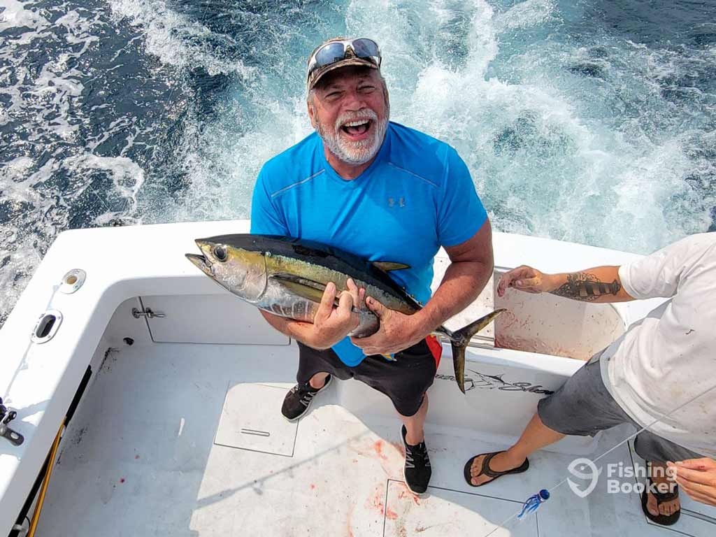 A proud angler in a blue shirt and white captain’s cap holds a smaller Tuna on a fishing boat during the offshore fishing trip in Virginia Beach