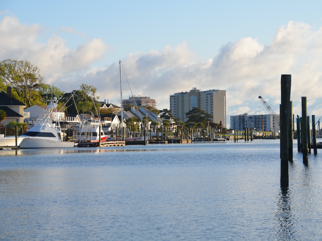 Boats docked at a marina, with waterfront houses, tall buildings, and a partly cloudy sky in the background.