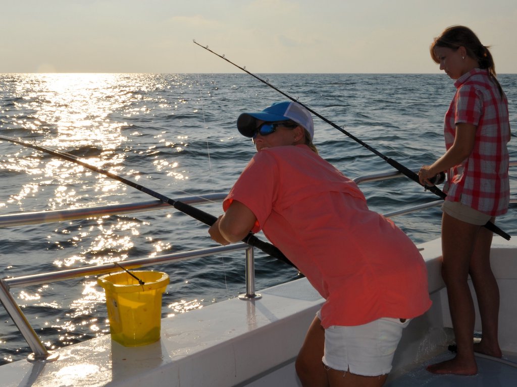 Two people are on a boat fishing in the ocean out of Virginia Beach during sunset, with a yellow bucket nearby and sunlight glinting off the water.