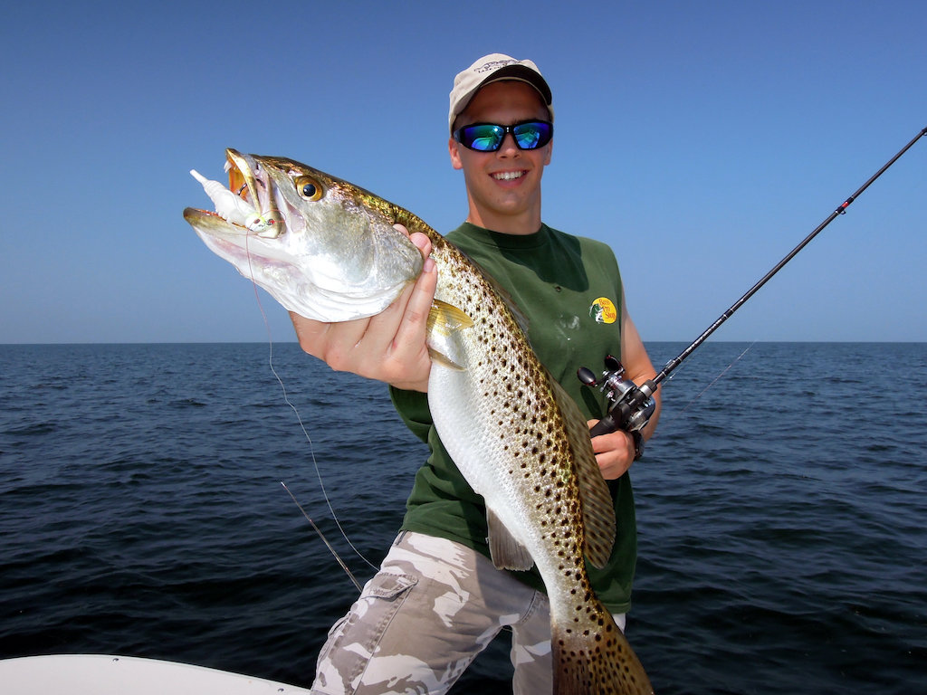 A man on a boat holding a large Speckled Trout fish with one hand and a fishing rod in the other.