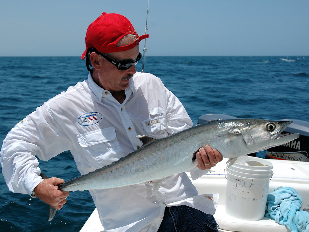 A man in a red cap and white shirt holding a large King Mackerel on a boat, with the ocean in the background.