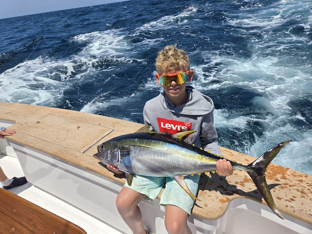 A boy in sunglasses and a grey Levi’s hoodie sits on a boat holding a large Yellowfin Tuna, with the ocean in the background.