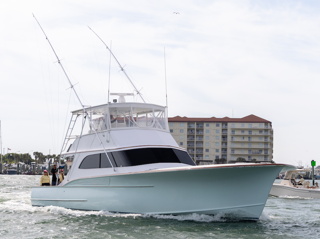 A white fishing yacht with several antennas moves through the water near a marina in Virginia Beach, with a multi-story building and other boats in the background.