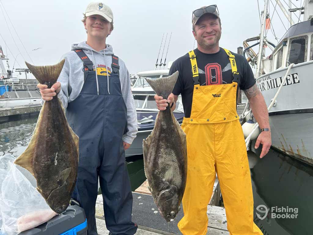 Two anglers in waterproof overalls stand on a dock in Washington holding decent Pacific Halibut, with boats and fishing gear in the background.