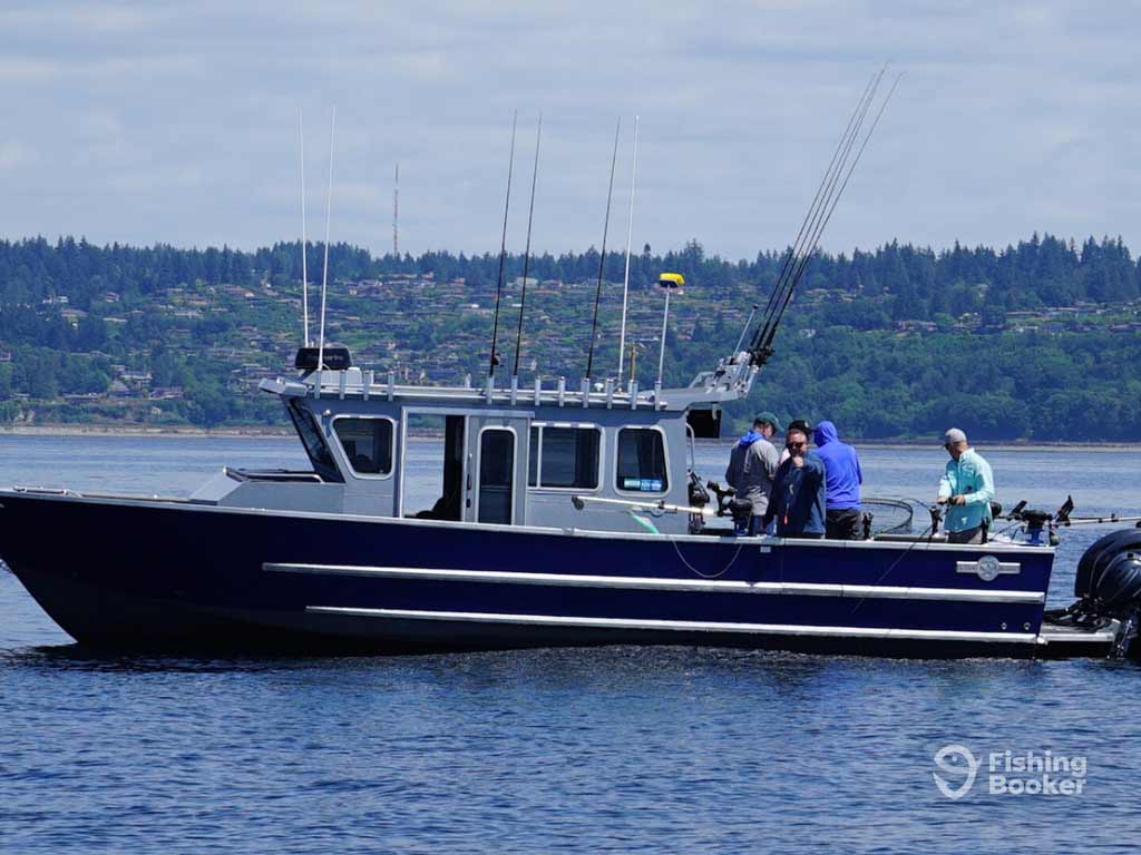 A group of anglers is fishing on a dark-colored boat with several rods, floating on calm water near a tree-lined shore.