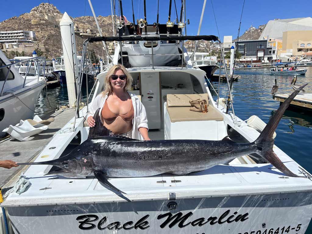 A woman on a boat at a marina poses with a large Black Marlin she caught during her Cabo San Lucas deep sea fishing trip, smiling in sunglasses under a clear blue sky.