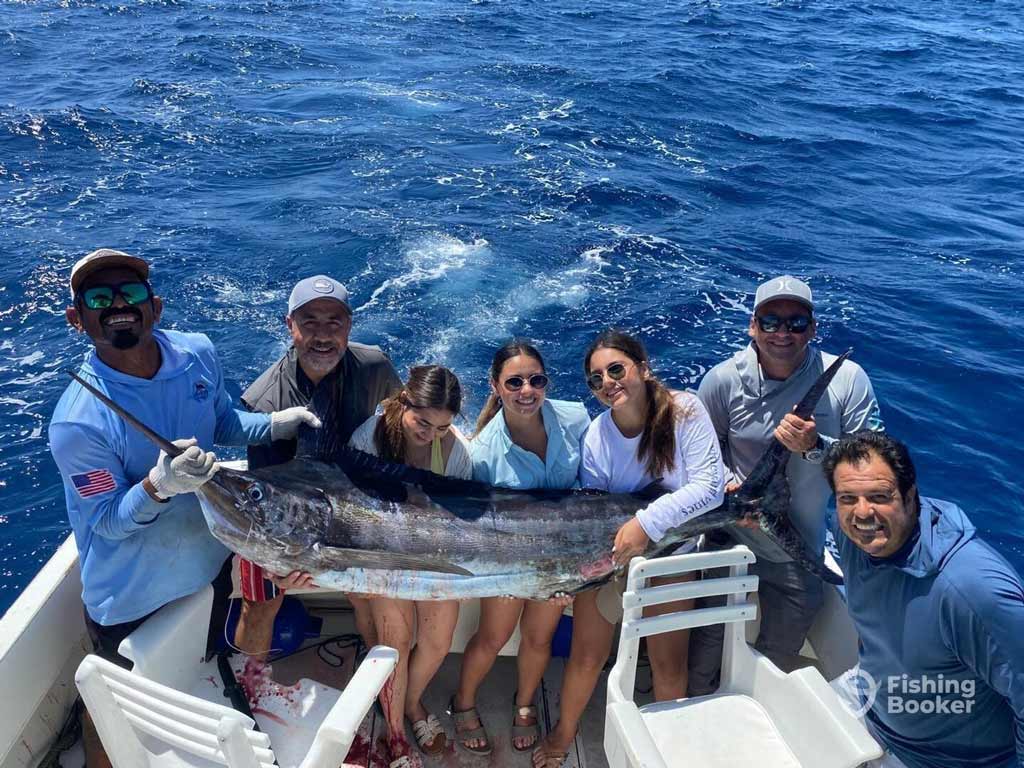 Seven anglers are posing on a charter boat while holding a large Marlin, a catch worthy of the Bisbees Black and Blue tournament. The Fishing Booker logo’s in the bottom right under the sunny sky.