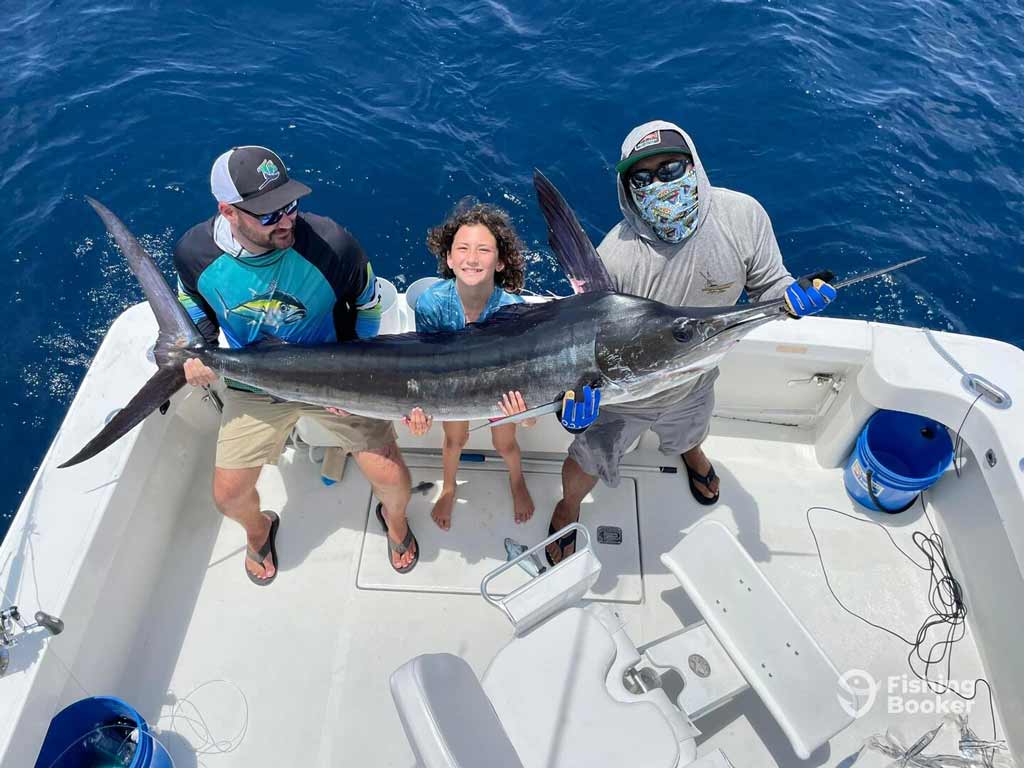 Three anglers standing on a boat and holding a large Marlin over blue ocean water in Cabo San Lucas - two adults and a child smile with fishing gear visible on the deck.