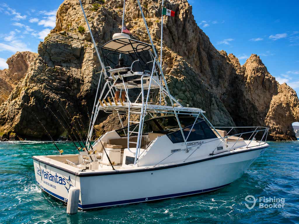 A white fishing boat floats on clear blue water near rocky cliffs, in Cabo San Lucas with rods mounted and a Mexican flag flying atop the boat.