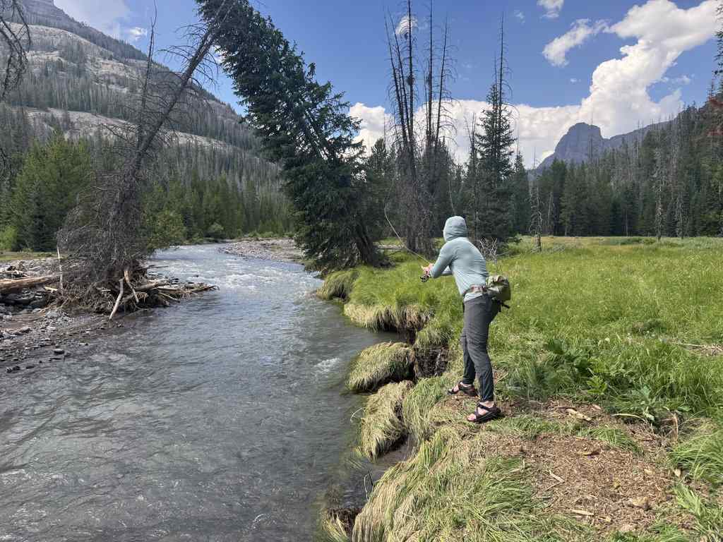 Someone’s wearing a hooded jacket and sandals, standing on a grassy riverbank as they cast a fishing line into the river, with trees and mountains in the background.