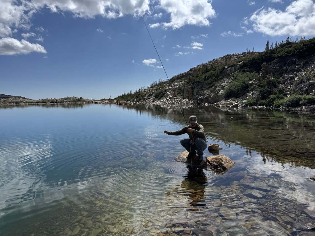 Someone’s fishing while kneeling on a rock in a clear, calm lake surrounded by rocky hills and sparse trees under a partly cloudy sky. No fish like Bass or Trout are visible.