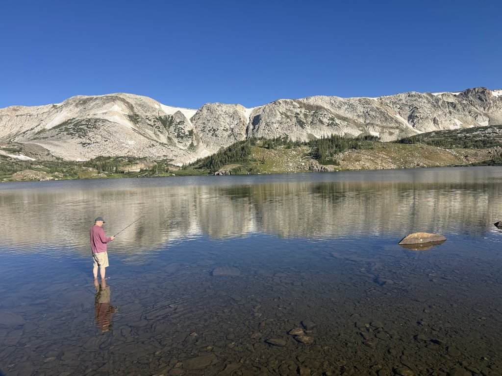Someone’s fishing while standing in shallow water in a clear lake, with a rocky shoreline and mountains behind them under a blue sky.