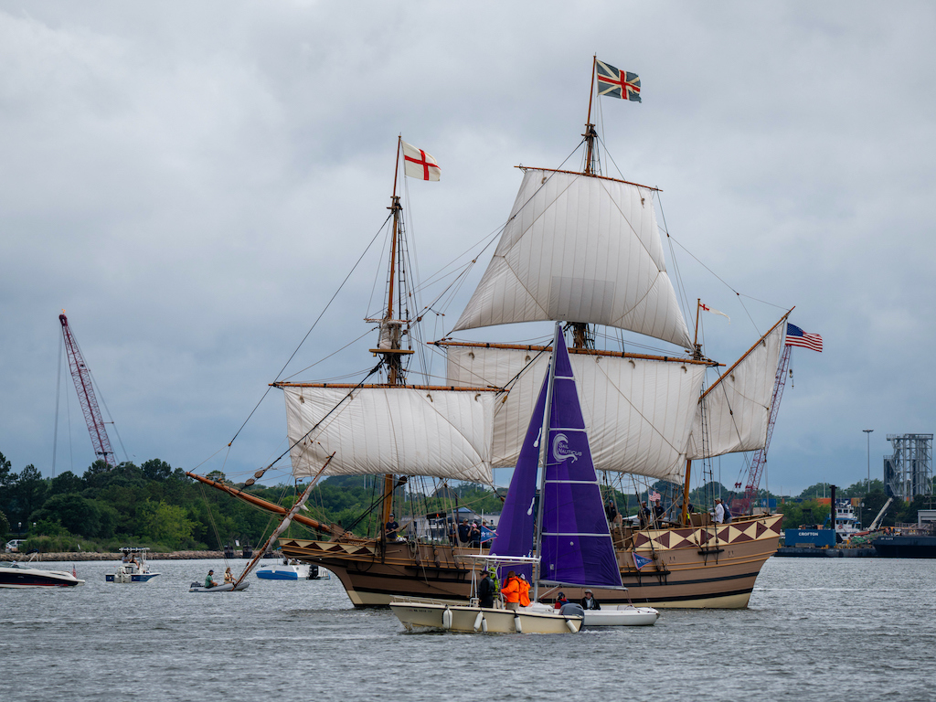 A large wooden sailing ship with tall masts and historic flags sails near Virginia Beach, surrounded by a small sailboat and other boats under a cloudy sky. 