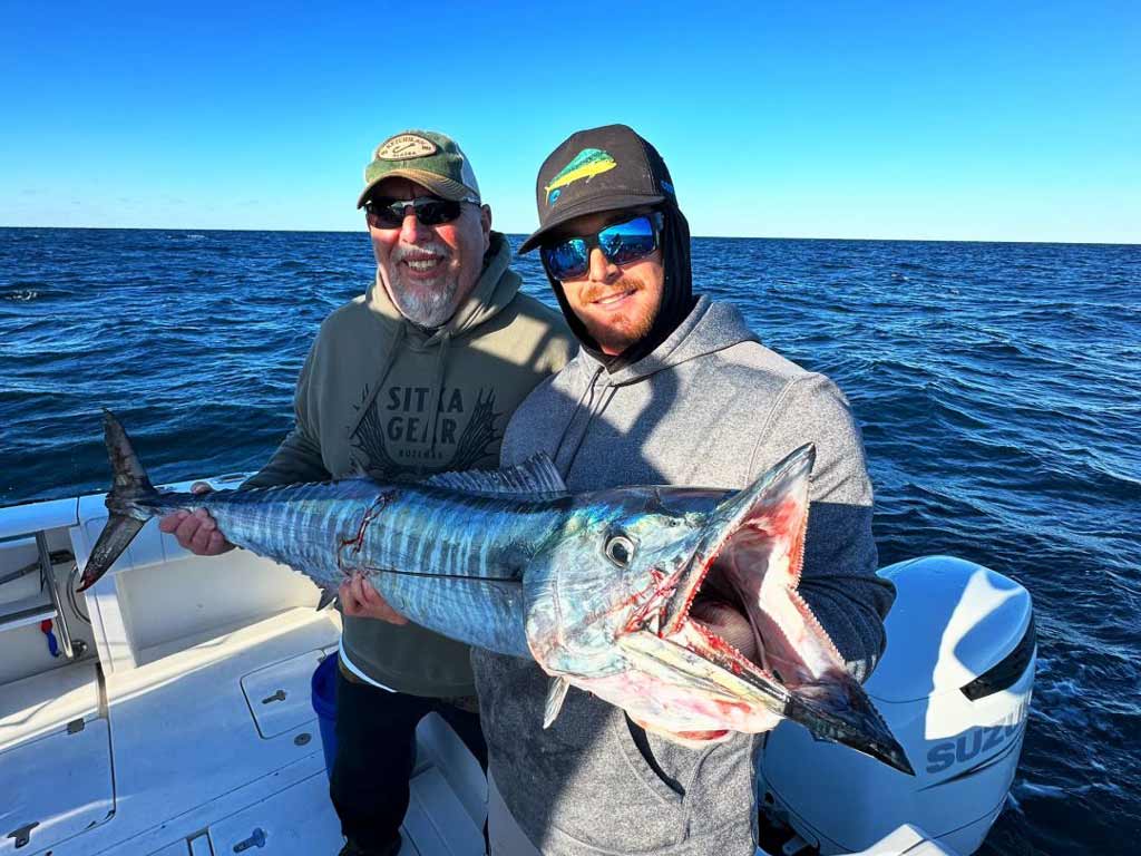 Two anglers standing on a charter boat and holding a large Wahoo over open water under a clear sky at Orange Beach, Alabama