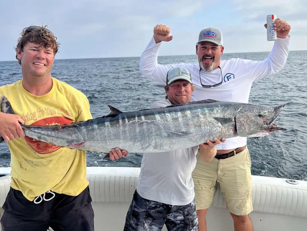 Three proud anglers are standing on a boat and holding a large Wahoo, a catch worthy of the Blue Marlin Grand Championship fishing tournament; ocean water glistens behind, while one man’s raising his arms and holding a can.