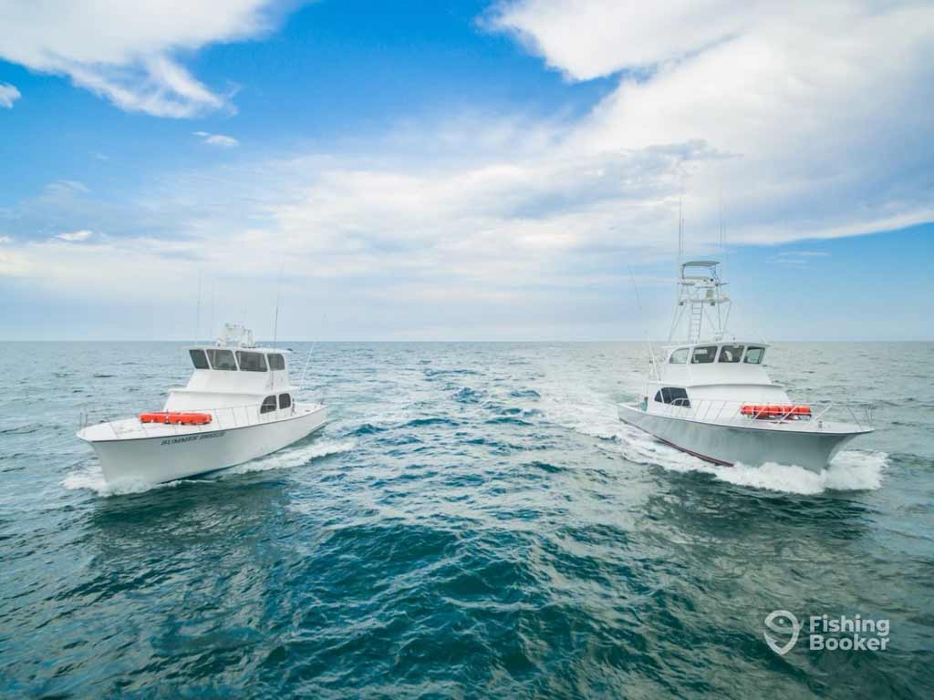 Two white sportfishing boats race side by side on open blue water under a partly cloudy sky, with the Fishing Booker logo in the corner.