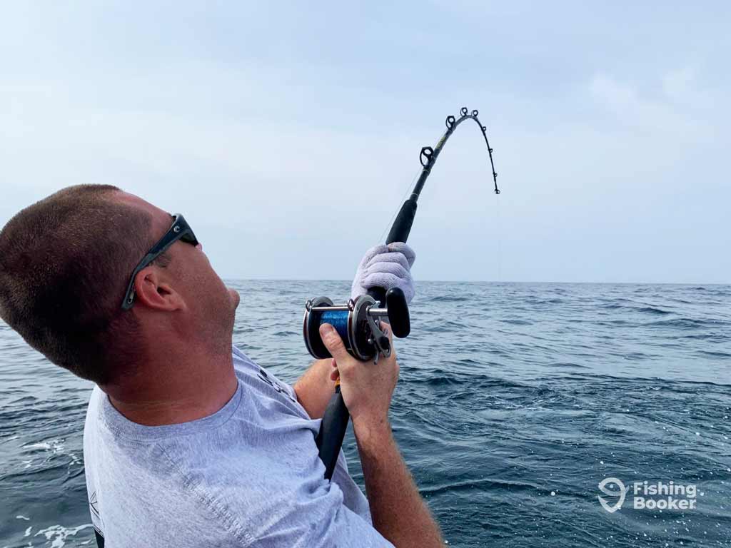 An angler is wearing sunglasses and gloves as they hold a bent fishing rod, struggling to reel in a massive gamefish, on the open sea beneath a cloudy Orange Beach sky.